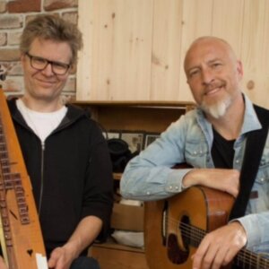 To menn med musikkinstrumenter, en har en gitar og den andre en langeleik. Brick vegg og trepanel i bakgrunnen.