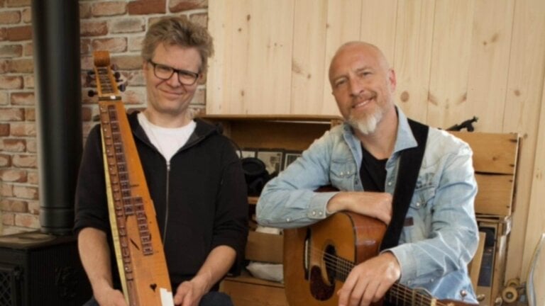 To menn med musikkinstrumenter, en har en gitar og den andre en langeleik. Brick vegg og trepanel i bakgrunnen.