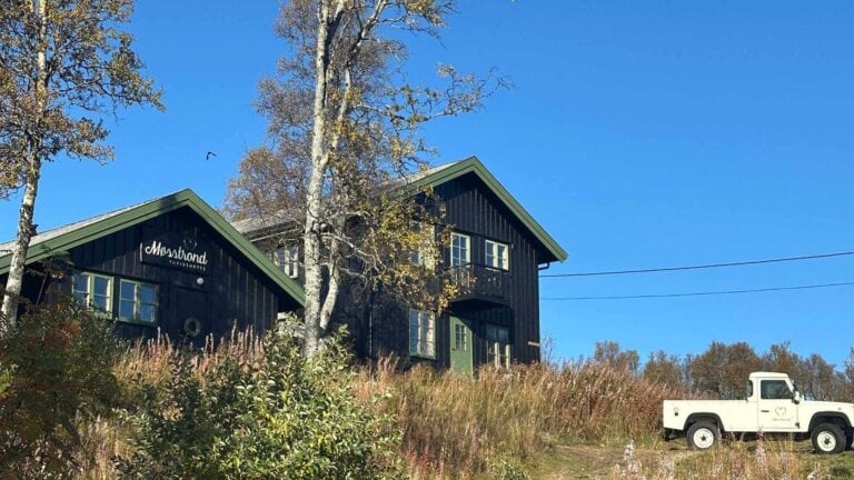 Svart trehytte, Mosstrond Turisthytte, med hvit bil i norsk natur på solrik dag.