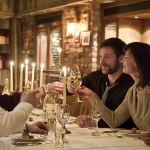Kvinner og menn skåler med vinglass i en koselig restaurant med levende lys.