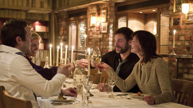 Kvinner og menn skåler med vinglass i en koselig restaurant med levende lys.