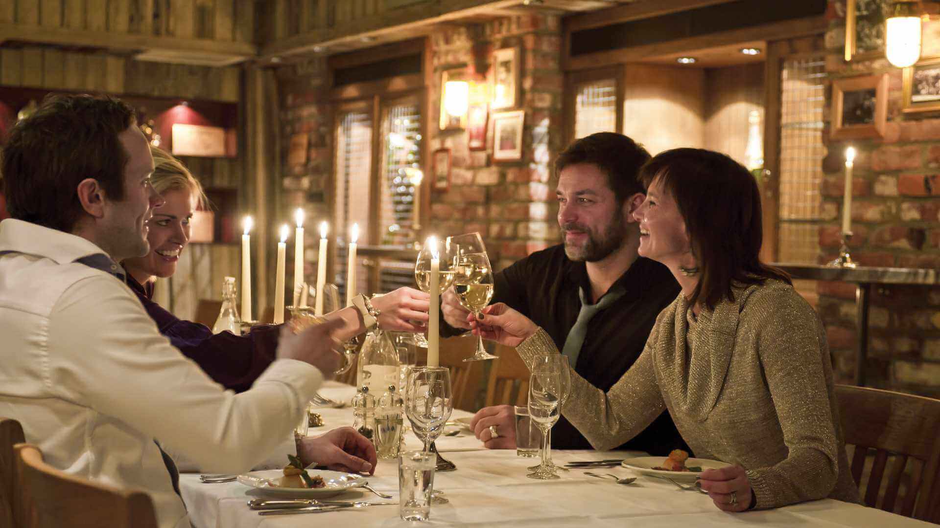 Kvinner og menn skåler med vinglass i en koselig restaurant med levende lys.