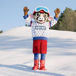 Maskot i vinterklær jubler på snødekt område med skog i bakgrunnen.