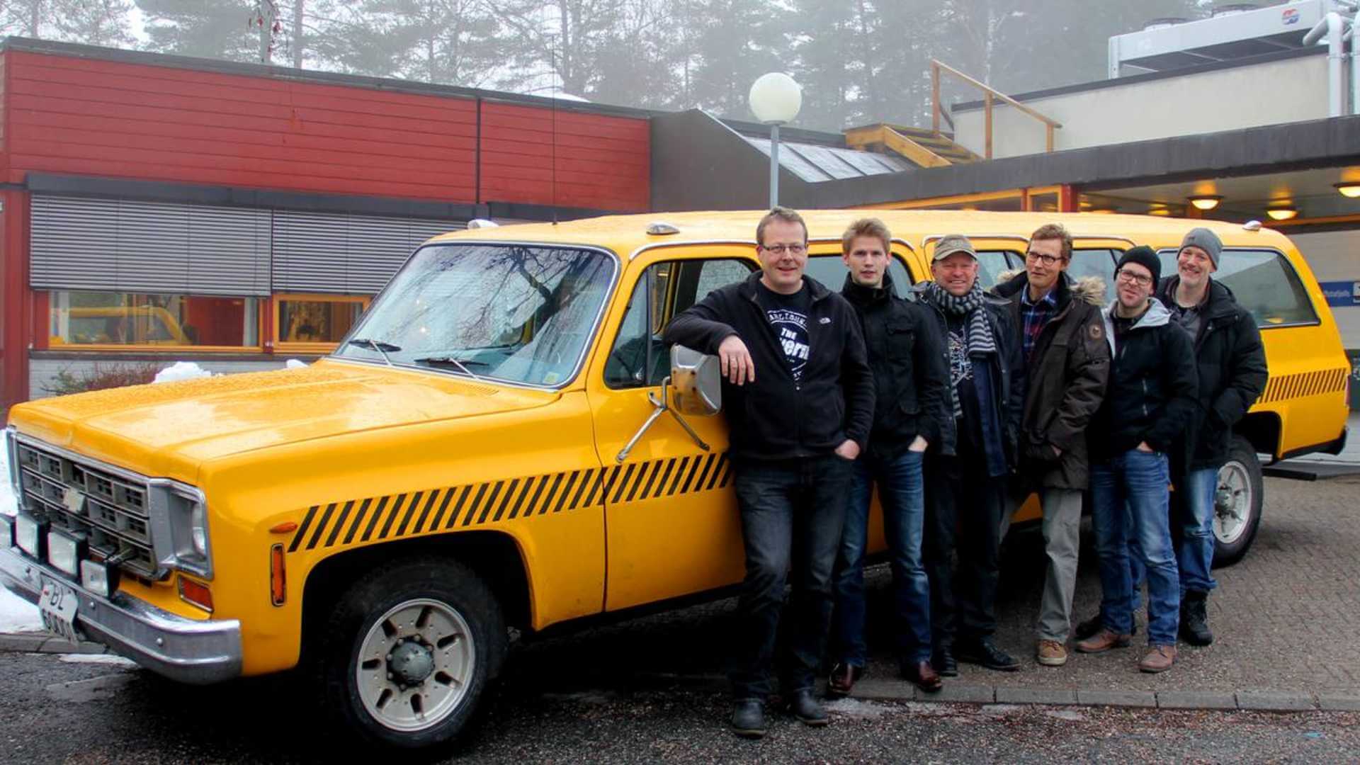 Gruppe menn foran gul varebil utenfor bygning på vinterdag.