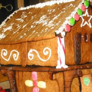 Pepperkakehus med fargerik pynt og hvit glasur på taket, plassert på et trebord nær et vindu.