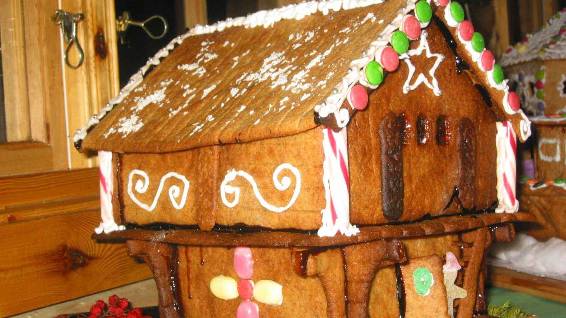 Pepperkakehus med fargerik pynt og hvit glasur på taket, plassert på et trebord nær et vindu.