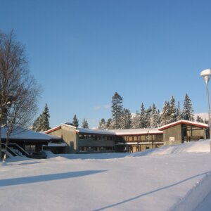 Snødekket skolebygning med klar blå himmel og skog i bakgrunnen, vinterstemning.