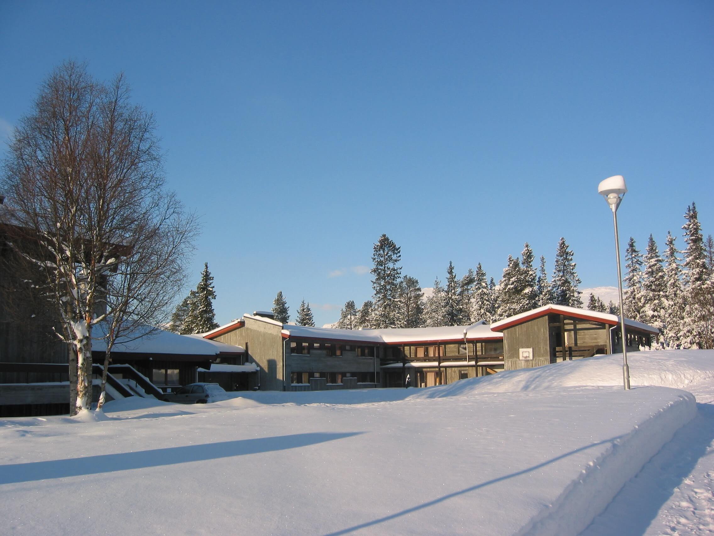 Snødekket skolebygning med klar blå himmel og skog i bakgrunnen, vinterstemning.