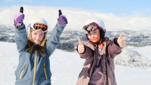 To barn i skiklær gir tommel opp i snødekte fjell. Klar himmel og vinterlandskap i bakgrunnen.