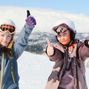 To barn i skiklær gir tommel opp i snødekte fjell. Klar himmel og vinterlandskap i bakgrunnen.