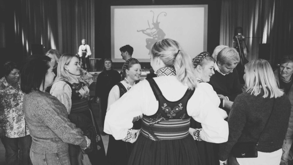 Eine Gruppe von Menschen in traditionellen norwegischen Trachten bei einem Treffen. Im Hintergrund ein Bildschirm.