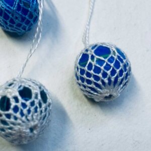 Blå perler i heklet netting, tre stykker, på hvit bakgrunn. Håndlagde dekorasjoner med delikat mønster.
