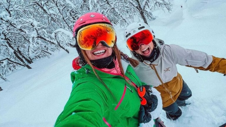 To glade skiløpere tar en selfie i et snødekket landskap med skiutstyr og hjelmer.
