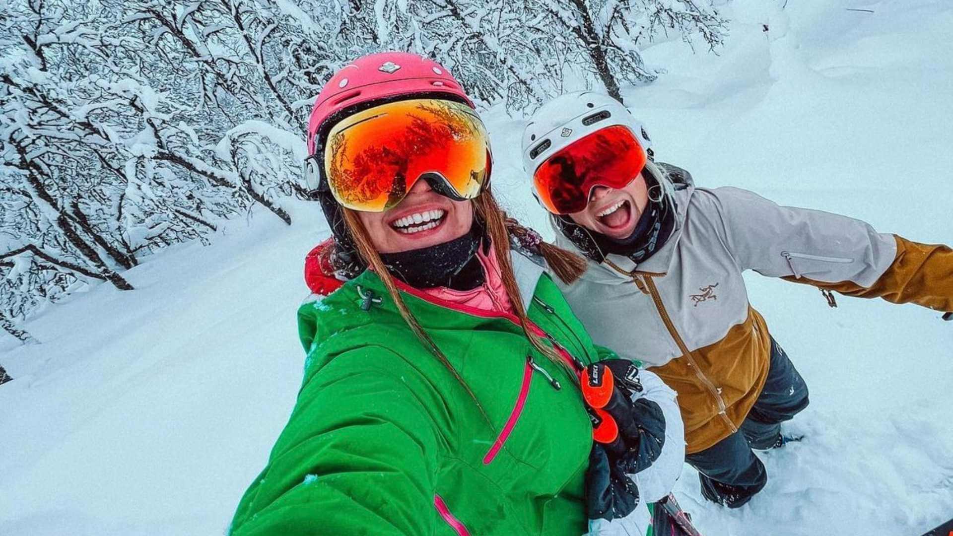 To glade skiløpere tar en selfie i et snødekket landskap med skiutstyr og hjelmer.