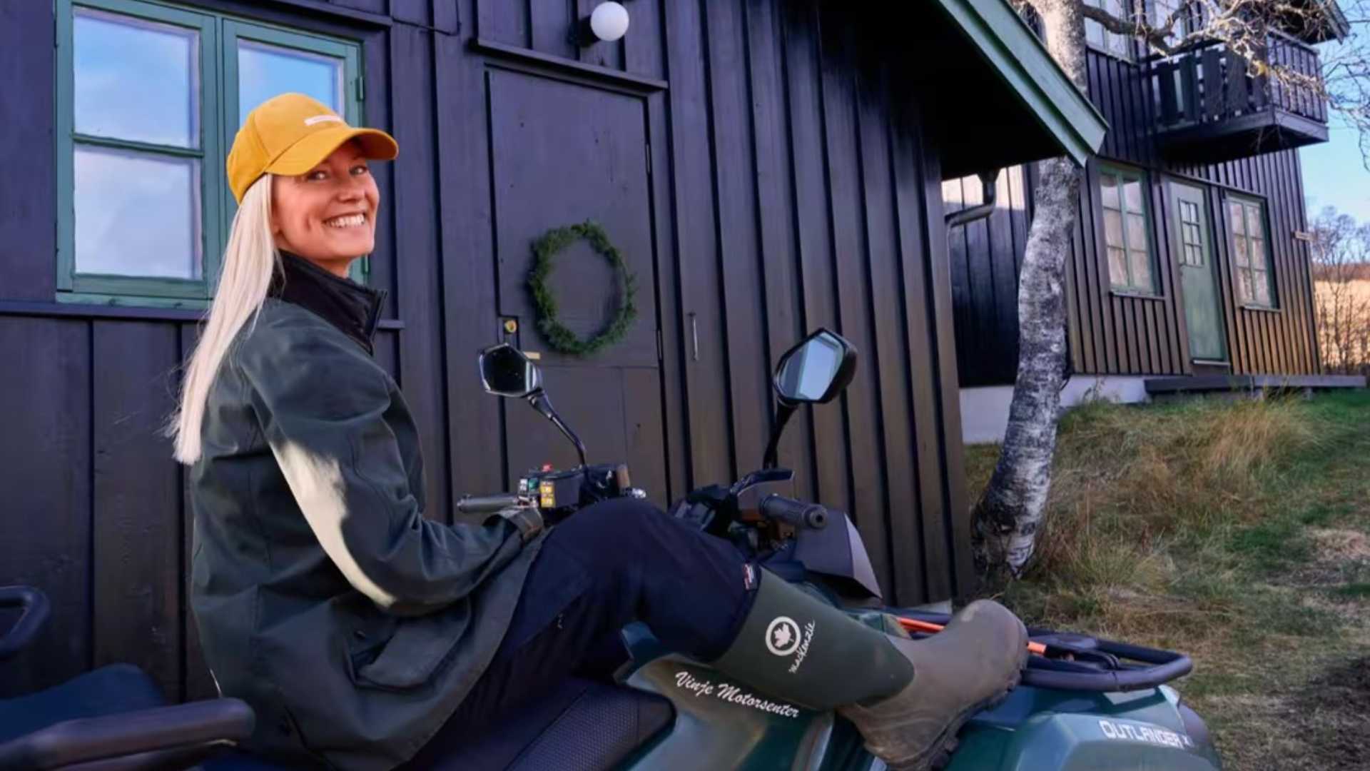 Kvinne i gul caps på ATV foran svart trehus med krans på døren, smiler ute i naturen.