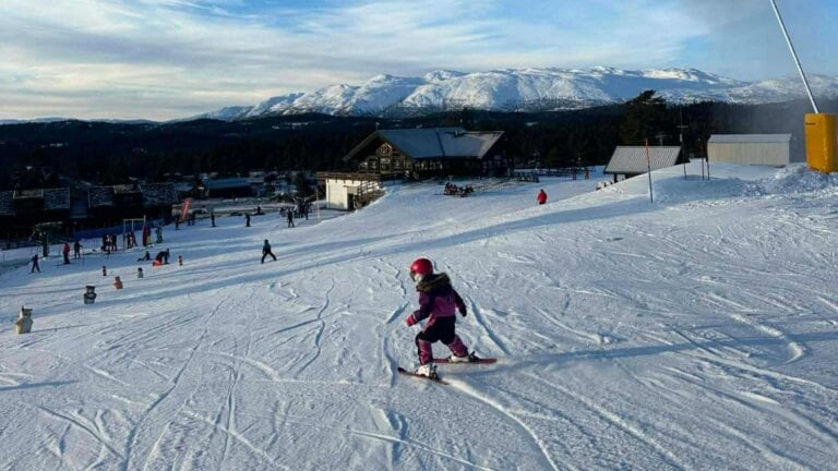 Barn står på ski i solfylt norsk fjellandskap, med snødekte fjell i bakgrunnen.