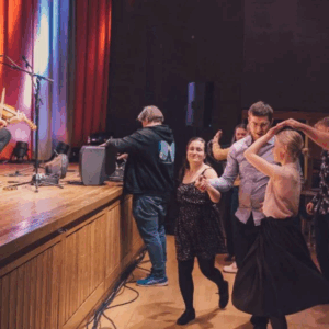 Musiker spiller fele på scenen mens folk danser i en sal. Levende kultur og folkemusikkopplevelse.