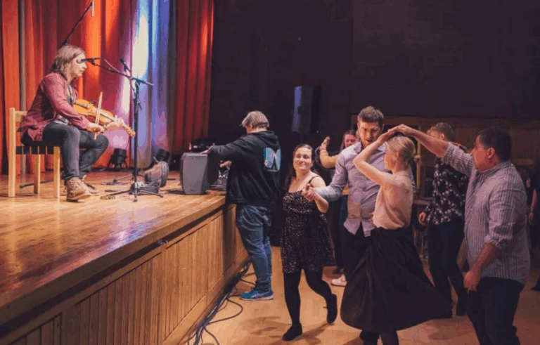 Musiker spiller fele på scenen mens folk danser i en sal. Levende kultur og folkemusikkopplevelse.