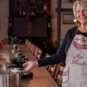 Kvinne i forkle på kjøkken, holder kasserolle ved et langt trebord i en koselig spisestue.