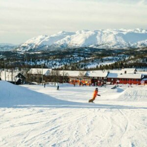 Snowboarder i et norsk skianlegg med snødekte fjell og trebygninger i bakgrunnen. Klar vinterhimmel.