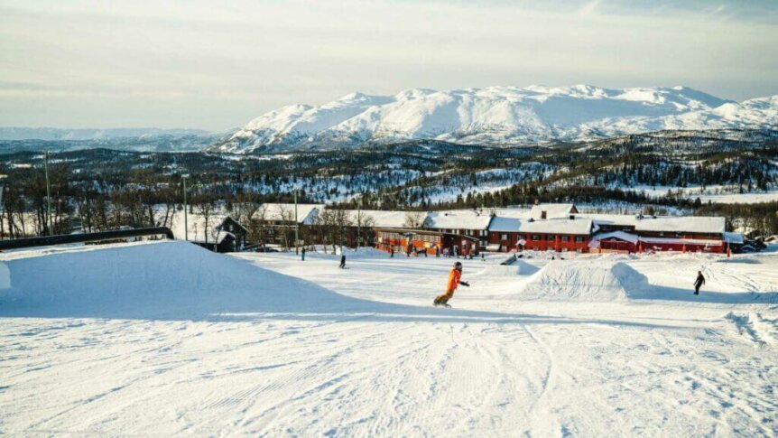 Snowboarder i et norsk skianlegg med snødekte fjell og trebygninger i bakgrunnen. Klar vinterhimmel.