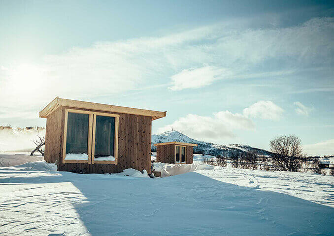 To små hytter i vinterlandskap med fjell i bakgrunnen og blå himmel.