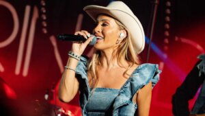 Kvinne med cowboyhatt synger på scenen med mikrofon, iført denimkjole, med rød belysning i bakgrunnen.