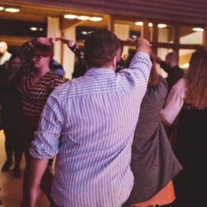 Folkedansere danser i en sal, kledd i tradisjonelle og moderne klær. Festlig sammenkomst i Norge.