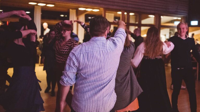 Folkedansere danser i en sal, kledd i tradisjonelle og moderne klær. Festlig sammenkomst i Norge.