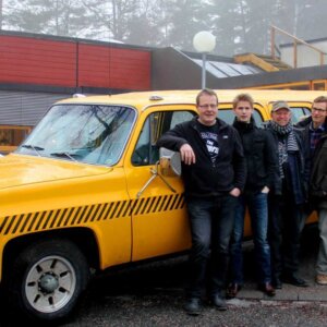 En gruppe menn poserer foran en gul vintage bil utenfor en bygning i vintermiljø.