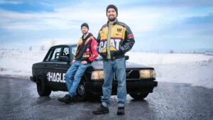 To personer i fargerike jakker poserer ved en svart bil om vinteren.