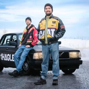 To personer i fargerike jakker poserer ved en svart bil om vinteren.