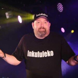 Mann på scenen med t-skjorte som sier Inkululeko og caps, i fargerikt opplyst konsertmiljø.