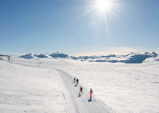 Langrennsløpere i solfylt snølandskap med fjell i bakgrunnen. Klar blå himmel og strålende vinterdag.