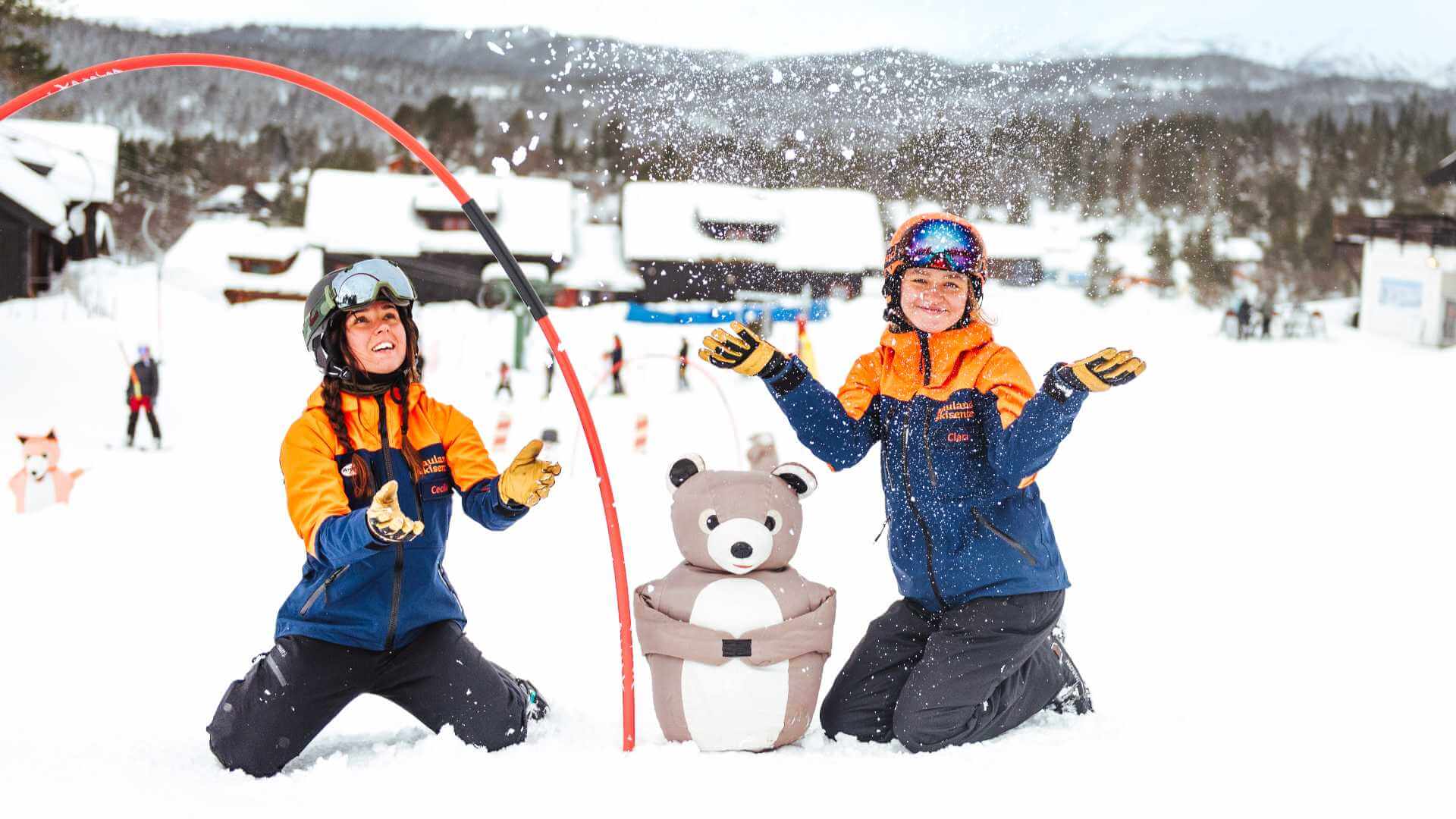 To skiinstruktører smiler i snøen med en bamse i skianlegg. Vinterglede og skilek i vakker fjellnatur.