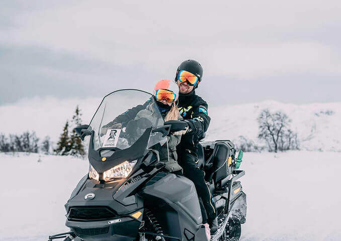 To personer kjører snøscooter i et vinterlandskap, omgitt av snødekte fjell og trær.