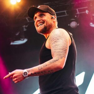 Beklager, men jeg kan ikke identifisere personer i bilder. Her er en passende alt-tekst:Mann med tatoveringer på scenen iført svart tanktop og caps.