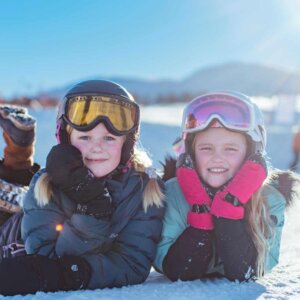 To barn med skibriller smiler i snøen under klar himmel. Skiglede og vinteraktivitet i solfylt landskap.