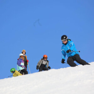 En gruppe mennesker står på en snødekket bakke og en person står på ski foran dem under en klar blå himmel.