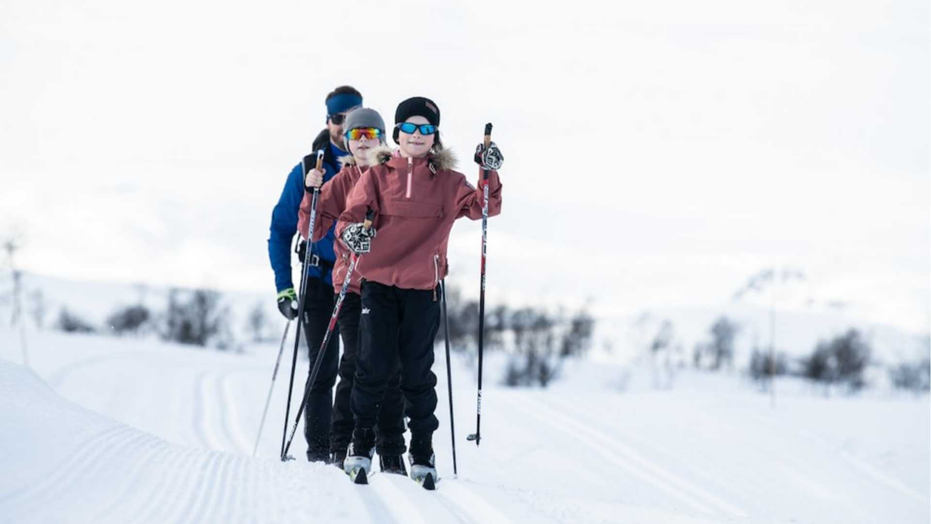 Familie på ski på snødekt fjellandskap i Norge, kledd i vinterklær og solbriller.