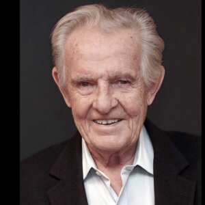 Eldre mann med grått hår og smilende ansikt, iført svart jakke og lys skjorte, foran mørk bakgrunn.