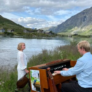 To mennesker ved elven, der en mann spiller på et piano. Vakker natur og gamle bygninger i bakgrunnen.