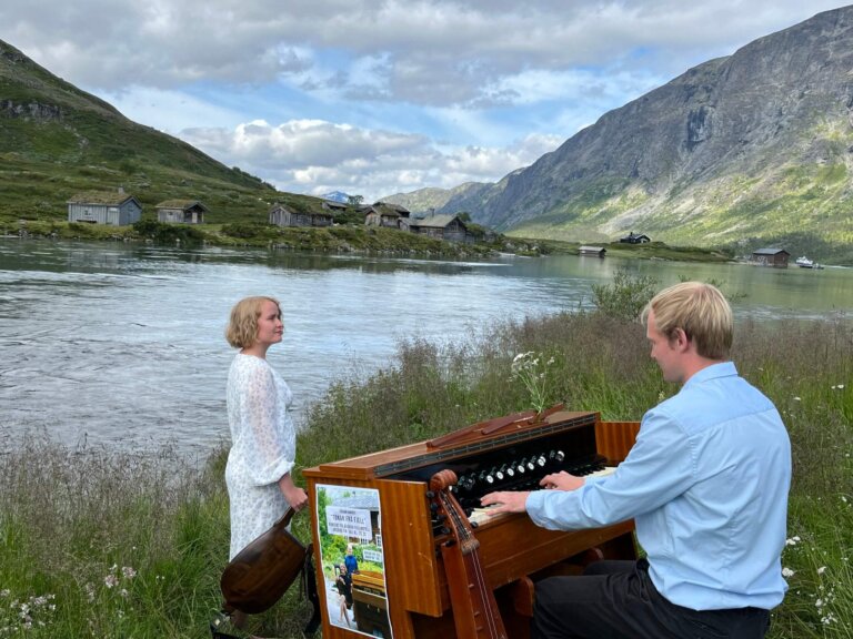 To mennesker ved elven, der en mann spiller på et piano. Vakker natur og gamle bygninger i bakgrunnen.