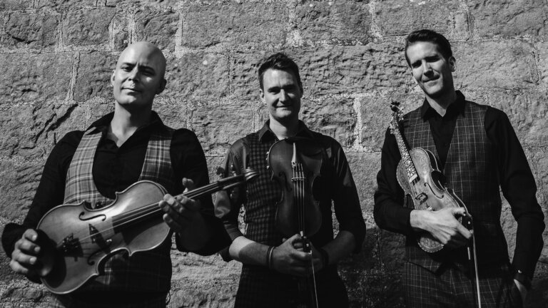 Tre musikere poserer med fioliner foran en steinvegg, iført stilige dresser, i svart-hvitt.