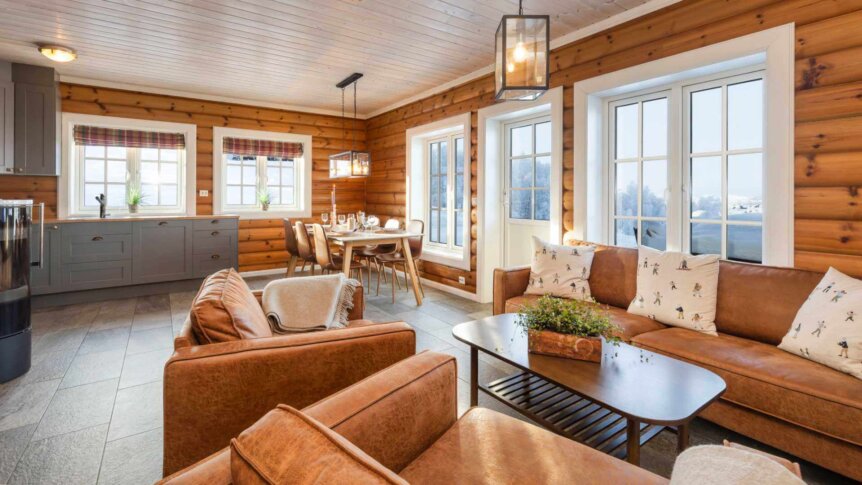 Koselig tømmerstue med brune skinnsofaer, spisestuebord og store vinduer med utsikt.