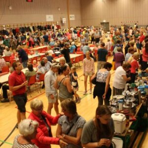Folkemasse på loppemarked med boder, salg av varer og mennesker som interagerer i en innendørs hall.