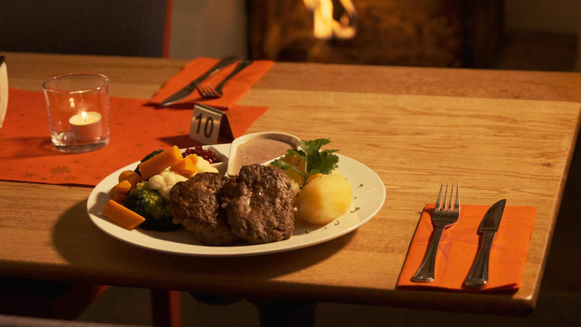 Hjemmelaget middag med karbonader, poteter, grønnsaker og saus på tallerken ved bord med orange serviett og lys.