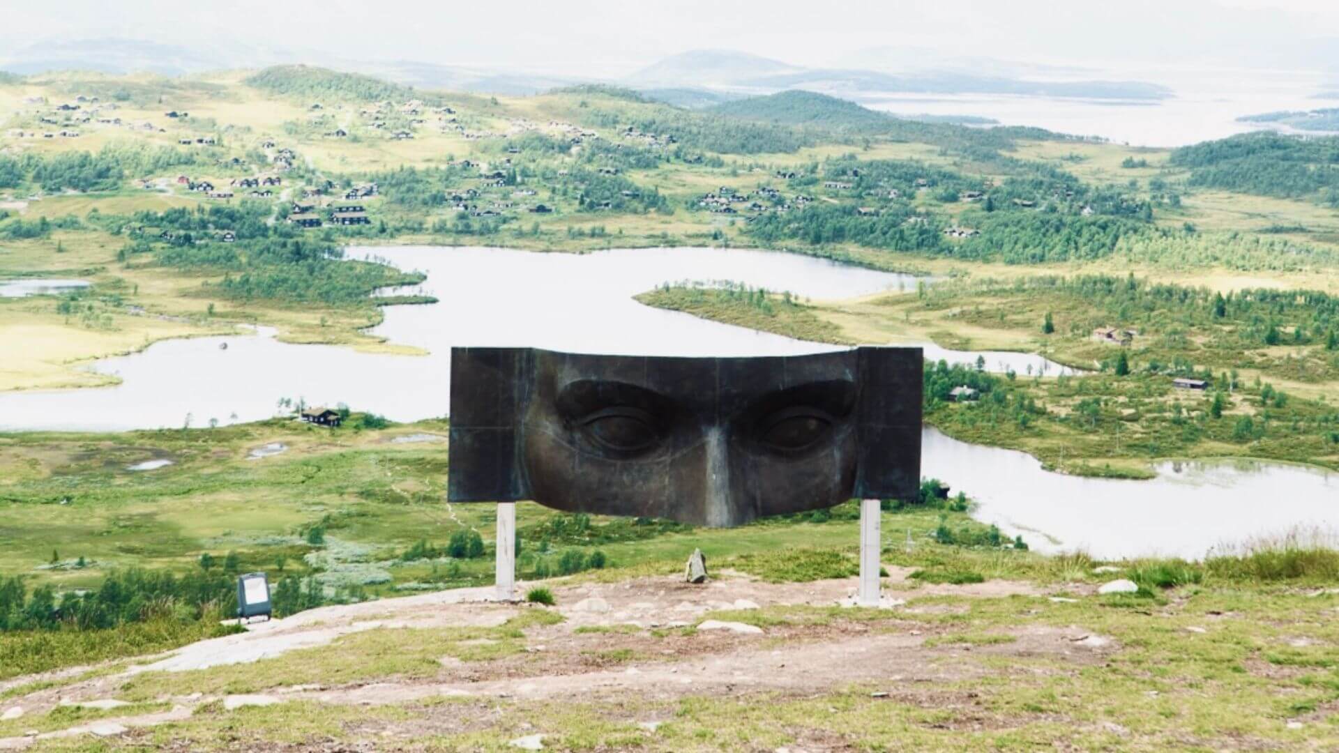 Storslått landskap med skulptur av et ansikt ved en innsjø, omkranset av fjell og grønn natur i Norge.