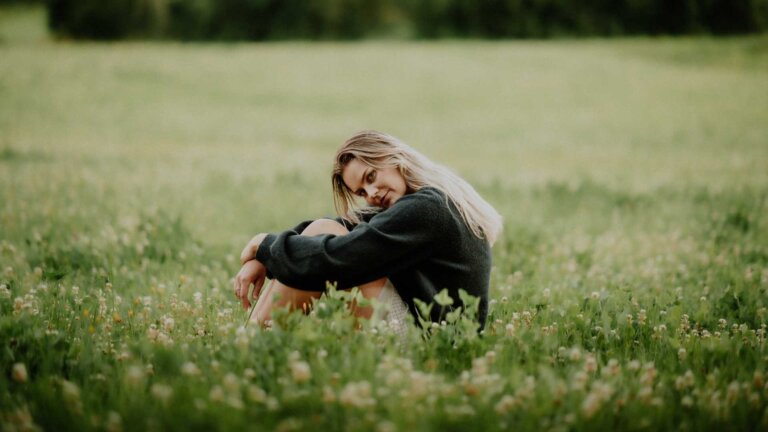 Ung kvinne sitter i en blomstereng, iført en mørk genser, med et tankefullt uttrykk.