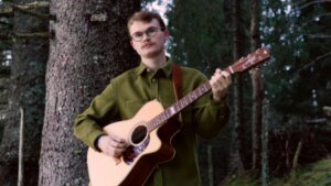 Ung mann med briller spiller gitar i skogen, kledd i grønn skjorte mot en bakgrunn av trær.